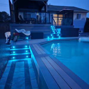 Above Ground Pool Deck with Upper Level Seating Area Pictured at Night With In-Deck Lighting Set to Light Blue.